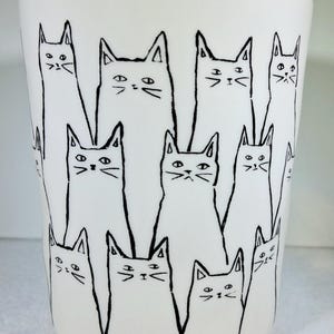 Puede incluir: Vaso de cer&aacute;mica blanco con un dibujo lineal negro repetido de gatos estilizados. Los gatos tienen orejas triangulares y rasgos faciales simples, dispuestos en un patr&oacute;n alrededor de la taza.