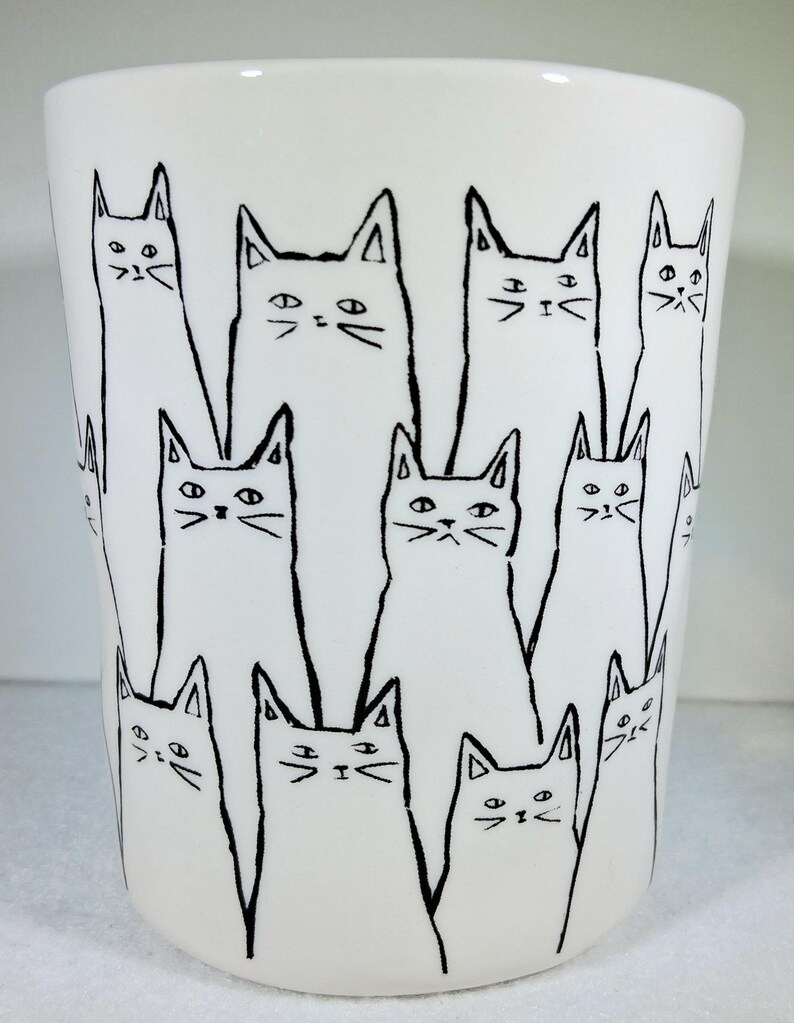 Puede incluir: Vaso de cer&aacute;mica blanco con un dibujo lineal negro repetido de gatos estilizados. Los gatos tienen orejas triangulares y rasgos faciales simples, dispuestos en un patr&oacute;n alrededor de la taza.