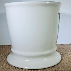 May include: A white ceramic tumbler with a flared base and a slightly tapered body. The tumbler has a simple design with a black letter on the side. The background is a neutral colour.