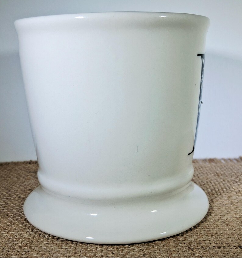 May include: A white ceramic tumbler with a flared base and a slightly tapered body. The tumbler has a simple design with a black letter on the side. The background is a neutral colour.