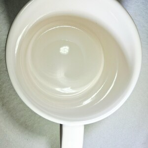May include: Overhead view of a white ceramic mug with a handle. The mug has a glossy finish and a simple, clean design. The interior of the mug is visible, showing a smooth, rounded bottom.