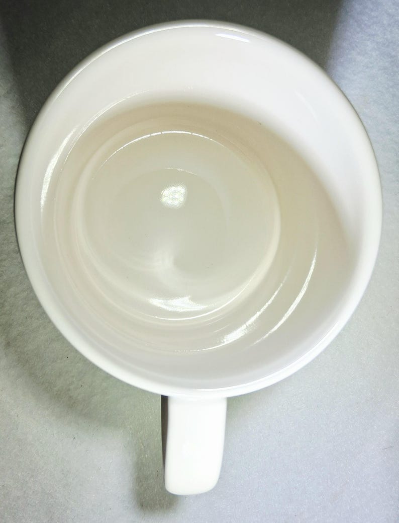 May include: Overhead view of a white ceramic mug with a handle. The mug has a glossy finish and a simple, clean design. The interior of the mug is visible, showing a smooth, rounded bottom.