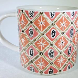 May include: White ceramic mug with a curved handle. The mug features a repeating pattern of pink, orange, and gold diamonds, with white starburst and black oval designs. The interior of the mug is white.