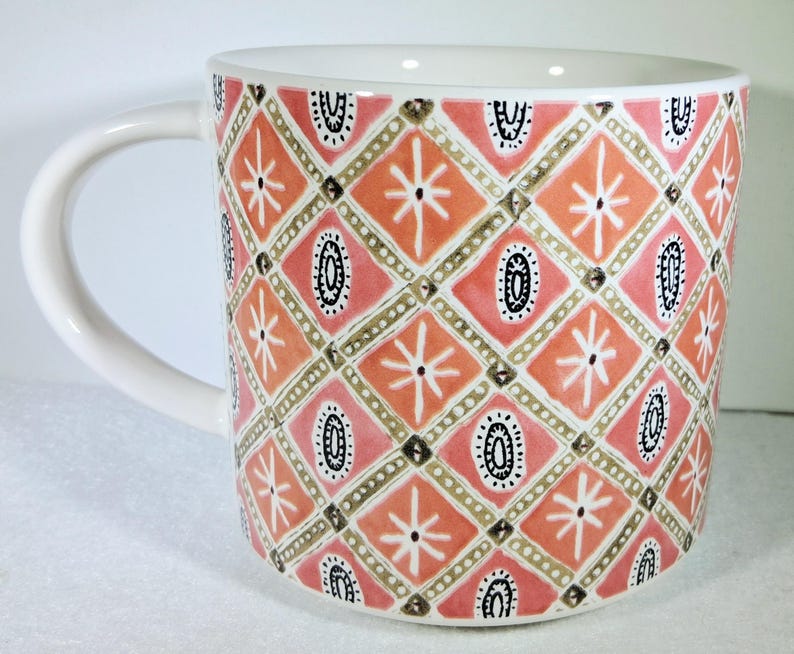 May include: White ceramic mug with a curved handle. The mug features a repeating pattern of pink, orange, and gold diamonds, with white starburst and black oval designs. The interior of the mug is white.