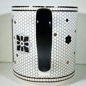 以下が含まれることがあります： 黒いハンドルと装飾的な模様が付いた白いセラミックマグ。マグカップは、黒と金のアクセントと黒い花のモチーフを備えたハニカムデザインが特徴です。リムとベースには、黒と白のボーダーがあります。