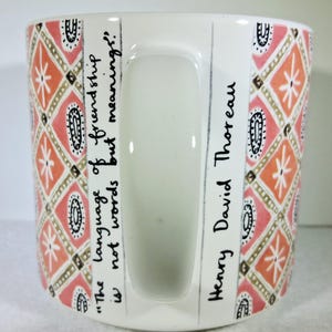 May include: White ceramic mug with a handle, featuring a repeating pattern of orange, pink, and gold geometric shapes. The mug has the quote "The language of friendship is not words but meanings!" and the name "Henry David Thoreau" written on it.