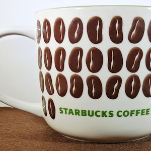 May include: White ceramic coffee mug with a repeating pattern of brown coffee beans. The mug has a white handle and the words "STARBUCKS COFFEE" in green lettering. The mug is on a brown surface.