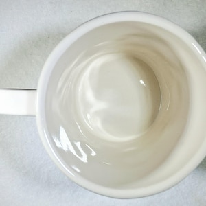Puede incluir: Vista superior de una taza de cer&aacute;mica blanca con asa. El interior de la taza es de color blanco cremoso y el asa es rectangular. La taza est&aacute; sobre una superficie blanca.