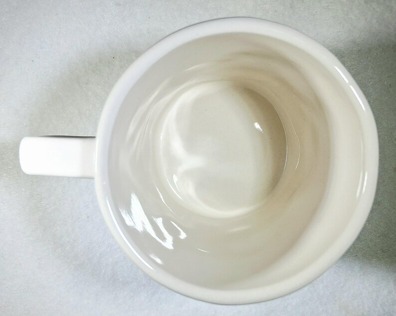 Puede incluir: Vista superior de una taza de cer&aacute;mica blanca con asa. El interior de la taza es de color blanco cremoso y el asa es rectangular. La taza est&aacute; sobre una superficie blanca.