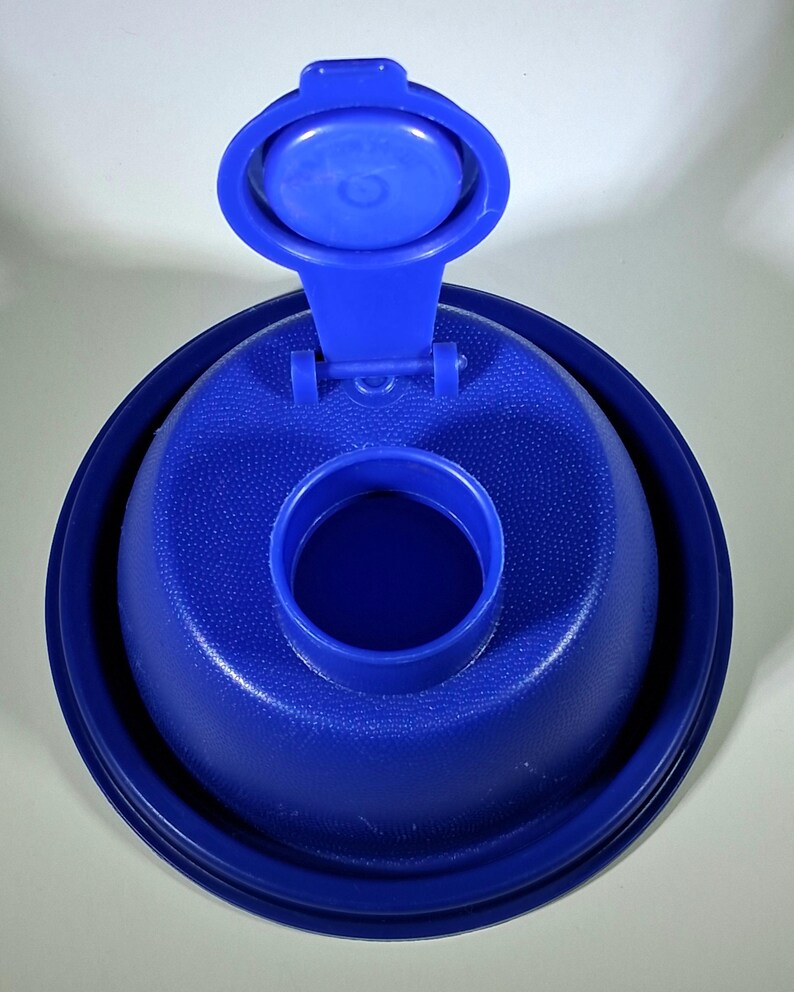 May include: A close-up of a blue plastic bottle cap with a hinged lid. The cap has a textured surface and a circular opening in the center. The lid is open, revealing the interior.