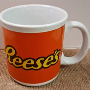 May include: Orange ceramic mug with the word "Reese's" in yellow and black lettering. The mug has a white interior and handle. The mug is a standard size for coffee or tea.