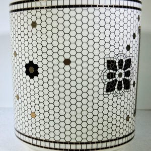 以下が含まれることがあります： 白地と白黒のハニカム模様の円筒形のセラミック容器。容器は黒と金のフローラルデザインと、上部と下部に黒いボーダーが特徴です。容器は装飾品です。