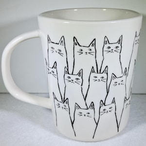 Puede incluir: Taza de cer&aacute;mica blanca con un dibujo lineal negro de varios gatos. Los gatos tienen orejas triangulares y rasgos faciales simples. La taza tiene un asa curva y un acabado liso y brillante.