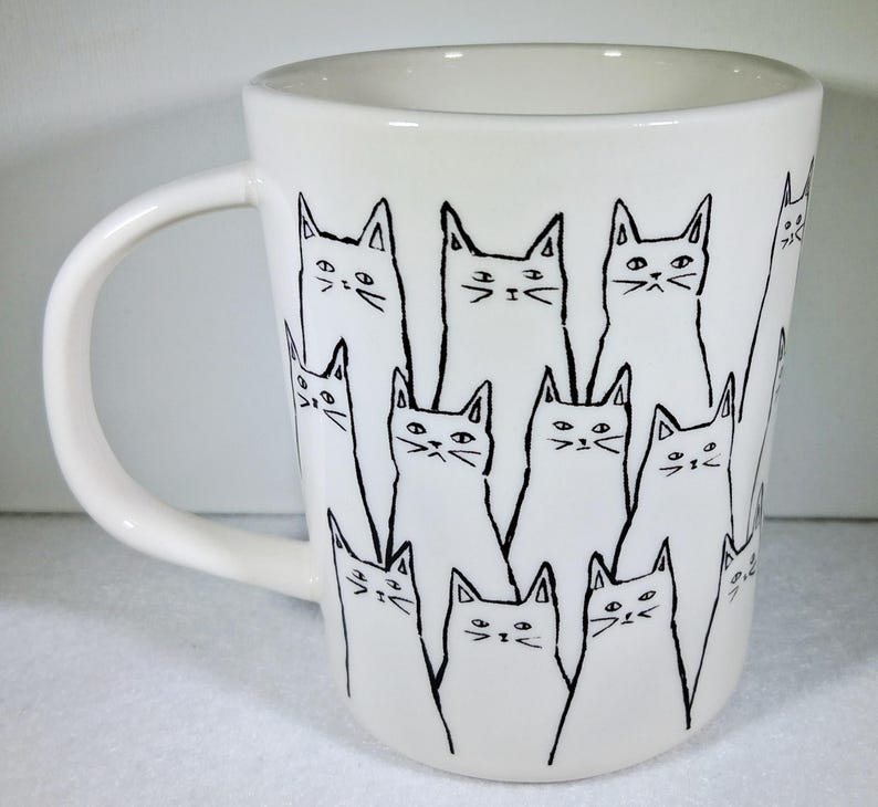 Puede incluir: Taza de cer&aacute;mica blanca con un dibujo lineal negro de varios gatos. Los gatos tienen orejas triangulares y rasgos faciales simples. La taza tiene un asa curva y un acabado liso y brillante.