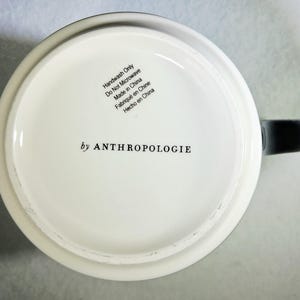 以下が含まれることがあります： 黒い持ち手が付いた白いセラミックマグ。マグの底には「by ANTHROPOLOGIE」の文字と、英語、フランス語、スペイン語によるお手入れ方法が記載されています。