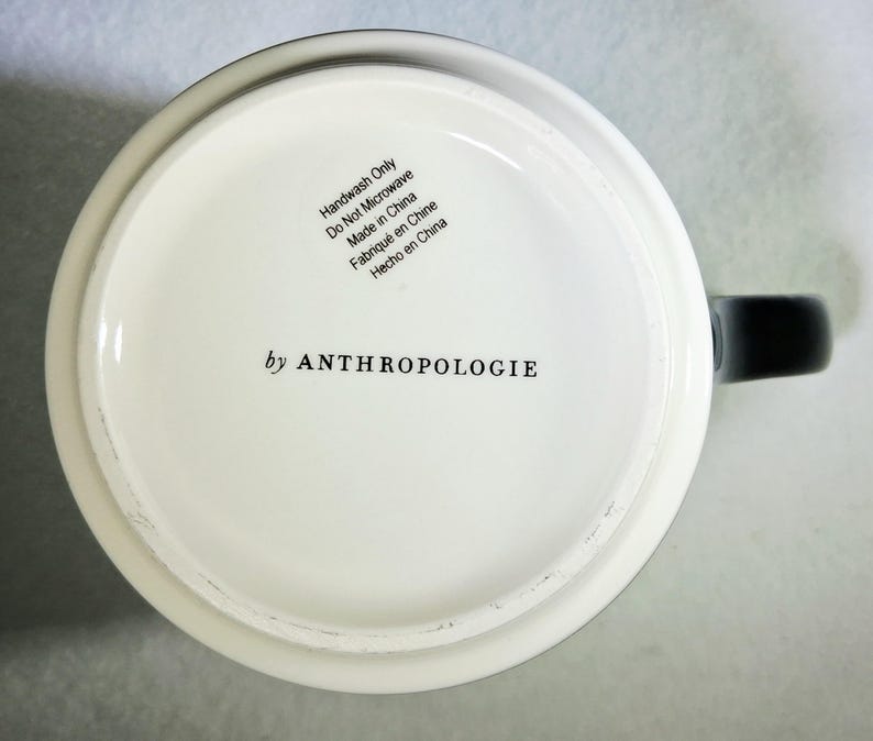 以下が含まれることがあります： 黒い持ち手が付いた白いセラミックマグ。マグの底には「by ANTHROPOLOGIE」の文字と、英語、フランス語、スペイン語によるお手入れ方法が記載されています。