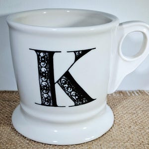 May include: White ceramic mug featuring a large black letter "K" with an ornate design. The mug has a rounded base and a handle. The design is detailed. The mug is a standard size.