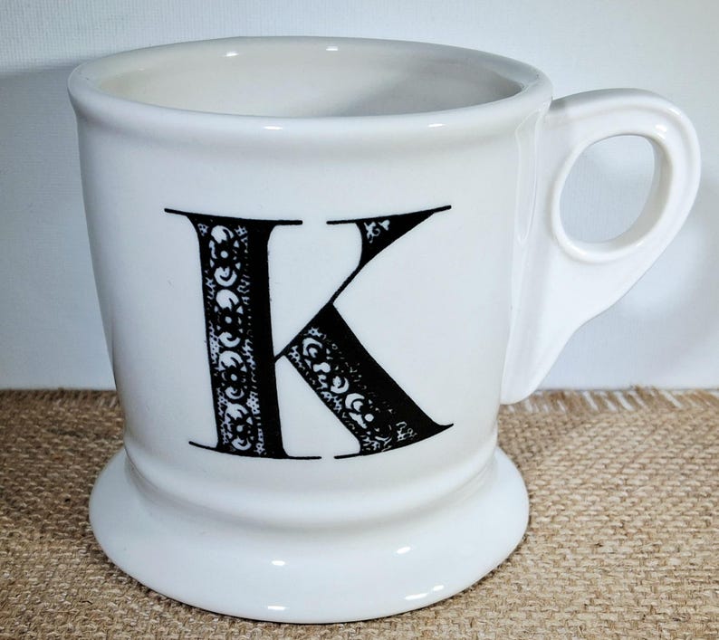May include: White ceramic mug featuring a large black letter "K" with an ornate design. The mug has a rounded base and a handle. The design is detailed. The mug is a standard size.