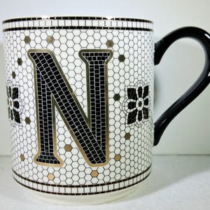 以下が含まれることがあります： 黒いハンドルが付いた白いセラミックマグ。黒と金の文字「N」のモザイクデザインが特徴です。マグカップは、黒と白の花柄と金のアクセントが付いています。