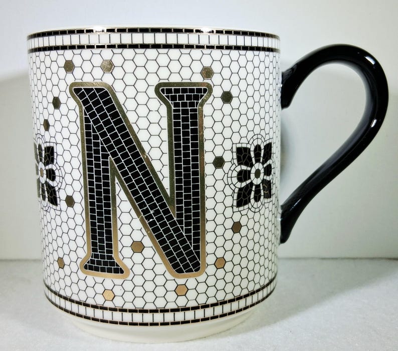 以下が含まれることがあります： 黒いハンドルが付いた白いセラミックマグ。黒と金の文字「N」のモザイクデザインが特徴です。マグカップは、黒と白の花柄と金のアクセントが付いています。