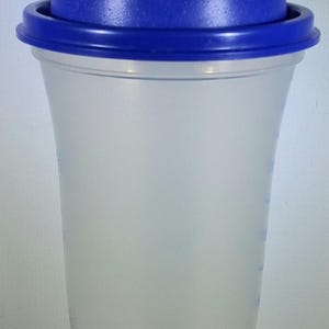 May include: A clear plastic shaker bottle with a blue lid and cap. The bottle has a wide base that tapers to a narrower top. The blue lid has a flip-top opening. The bottle has measurement markings on the side.