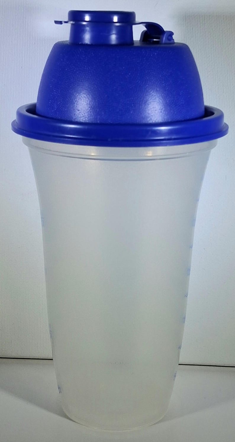 May include: A clear plastic shaker bottle with a blue lid and cap. The bottle has a wide base that tapers to a narrower top. The blue lid has a flip-top opening. The bottle has measurement markings on the side.