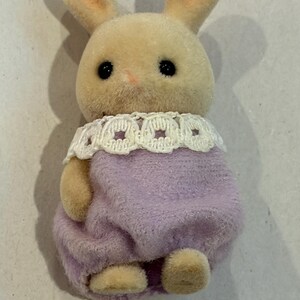 K&ouml;nnte beinhalten: Kleine, pl&uuml;schige Hasenfigur mit hellbraunem K&ouml;rper, langen Ohren und schwarzen Augen. Sie tr&auml;gt ein lavendelfarbenes Outfit mit einem wei&szlig;en Spitzenkragen. Das Spielzeug ist als Hase gestaltet.