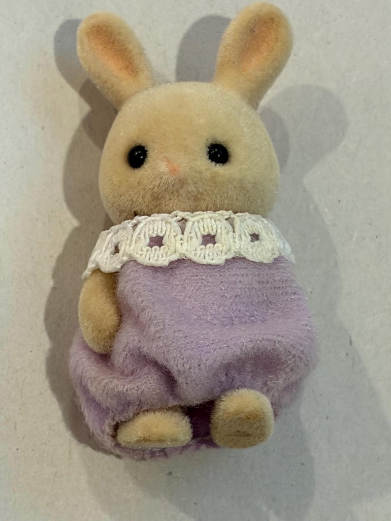 K&ouml;nnte beinhalten: Kleine, pl&uuml;schige Hasenfigur mit hellbraunem K&ouml;rper, langen Ohren und schwarzen Augen. Sie tr&auml;gt ein lavendelfarbenes Outfit mit einem wei&szlig;en Spitzenkragen. Das Spielzeug ist als Hase gestaltet.