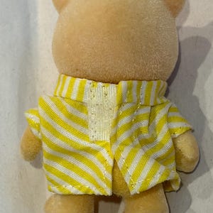 K&ouml;nnte beinhalten: Eine kleine, hellgelbe B&auml;renfigur mit einer gelb-wei&szlig; gestreiften Jacke. Die Jacke ist vorne offen und gibt einen Blick auf ein wei&szlig;es, glitzerndes Material frei. Das Fell des B&auml;ren ist weich und flauschig, mit kleinen, abgerundeten Ohren.