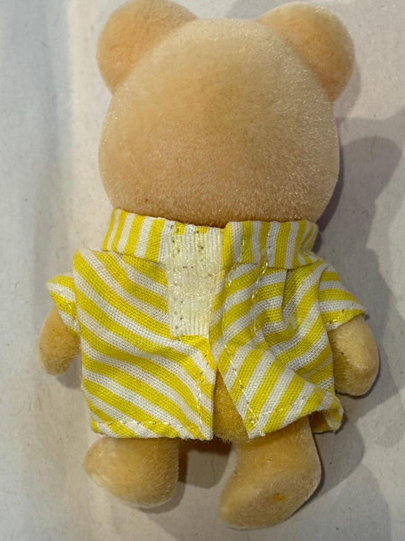 K&ouml;nnte beinhalten: Eine kleine, hellgelbe B&auml;renfigur mit einer gelb-wei&szlig; gestreiften Jacke. Die Jacke ist vorne offen und gibt einen Blick auf ein wei&szlig;es, glitzerndes Material frei. Das Fell des B&auml;ren ist weich und flauschig, mit kleinen, abgerundeten Ohren.