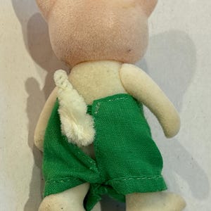 K&ouml;nnte beinhalten: Vintage Schweinchen-Figur, von hinten betrachtet, mit hellrosa Kopf und Ohren, cremefarbenem K&ouml;rper und gr&uuml;ner Hose. Das Schwein tr&auml;gt wei&szlig;e Schuhe und hat einen wei&szlig;en, flauschigen Schwanz.