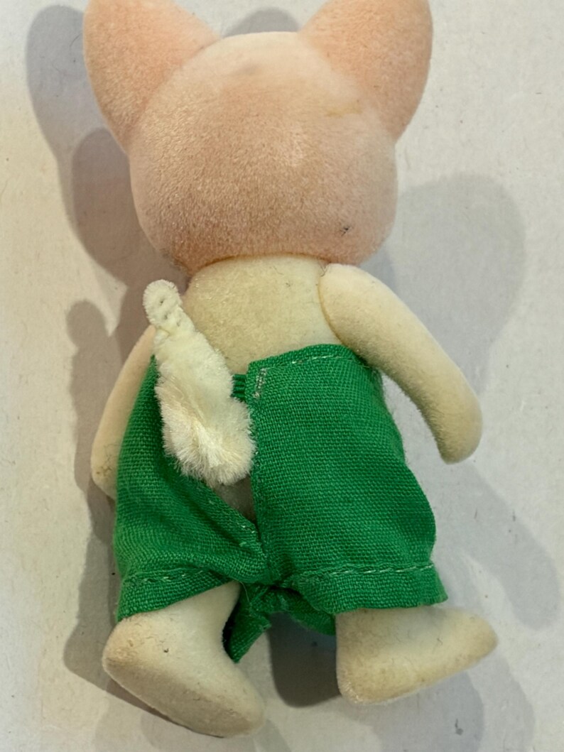 K&ouml;nnte beinhalten: Vintage Schweinchen-Figur, von hinten betrachtet, mit hellrosa Kopf und Ohren, cremefarbenem K&ouml;rper und gr&uuml;ner Hose. Das Schwein tr&auml;gt wei&szlig;e Schuhe und hat einen wei&szlig;en, flauschigen Schwanz.