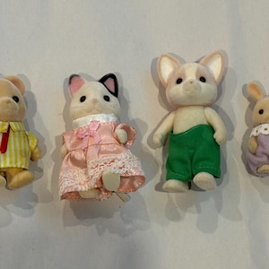 K&ouml;nnte beinhalten: Vier Miniatur-Tierfiguren: ein B&auml;r in einem gelb gestreiften Hemd, eine Katze in einem rosa Kleid, ein Hund in gr&uuml;nen Hosen und ein Kaninchen in einem lila Kleid. Diese Spielzeuge sind mit weichen Texturen und Details gestaltet.