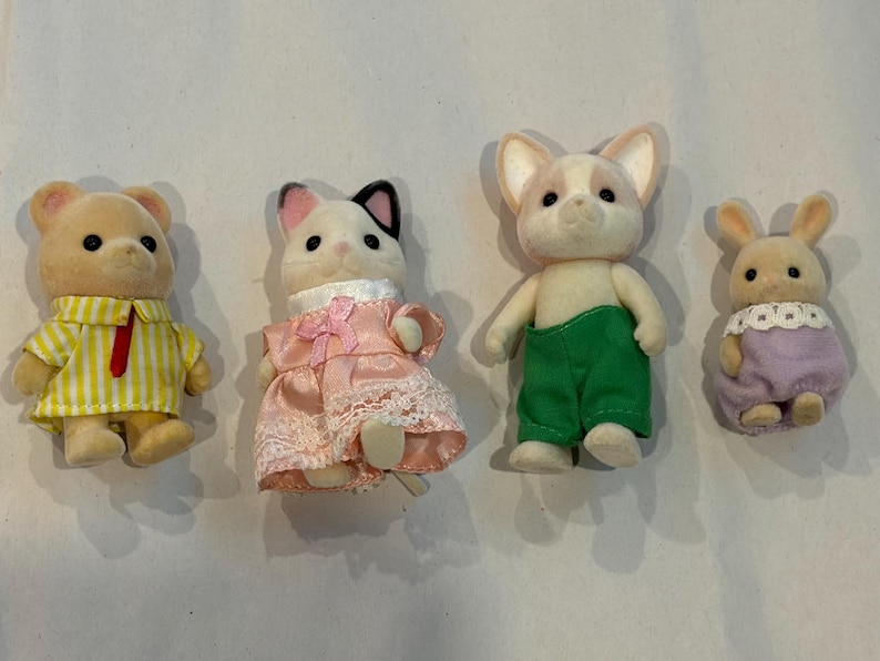 K&ouml;nnte beinhalten: Vier Miniatur-Tierfiguren: ein B&auml;r in einem gelb gestreiften Hemd, eine Katze in einem rosa Kleid, ein Hund in gr&uuml;nen Hosen und ein Kaninchen in einem lila Kleid. Diese Spielzeuge sind mit weichen Texturen und Details gestaltet.