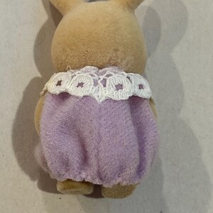K&ouml;nnte beinhalten: Eine hellbraune Spielzeug-Kaninchenfigur, von hinten gesehen, mit einem lavendelfarbenen Outfit und einem wei&szlig;en Spitzenkragen. Das Kaninchen hat lange Ohren und eine weiche, flauschige Textur, typisch f&uuml;r Kinderspielzeug.