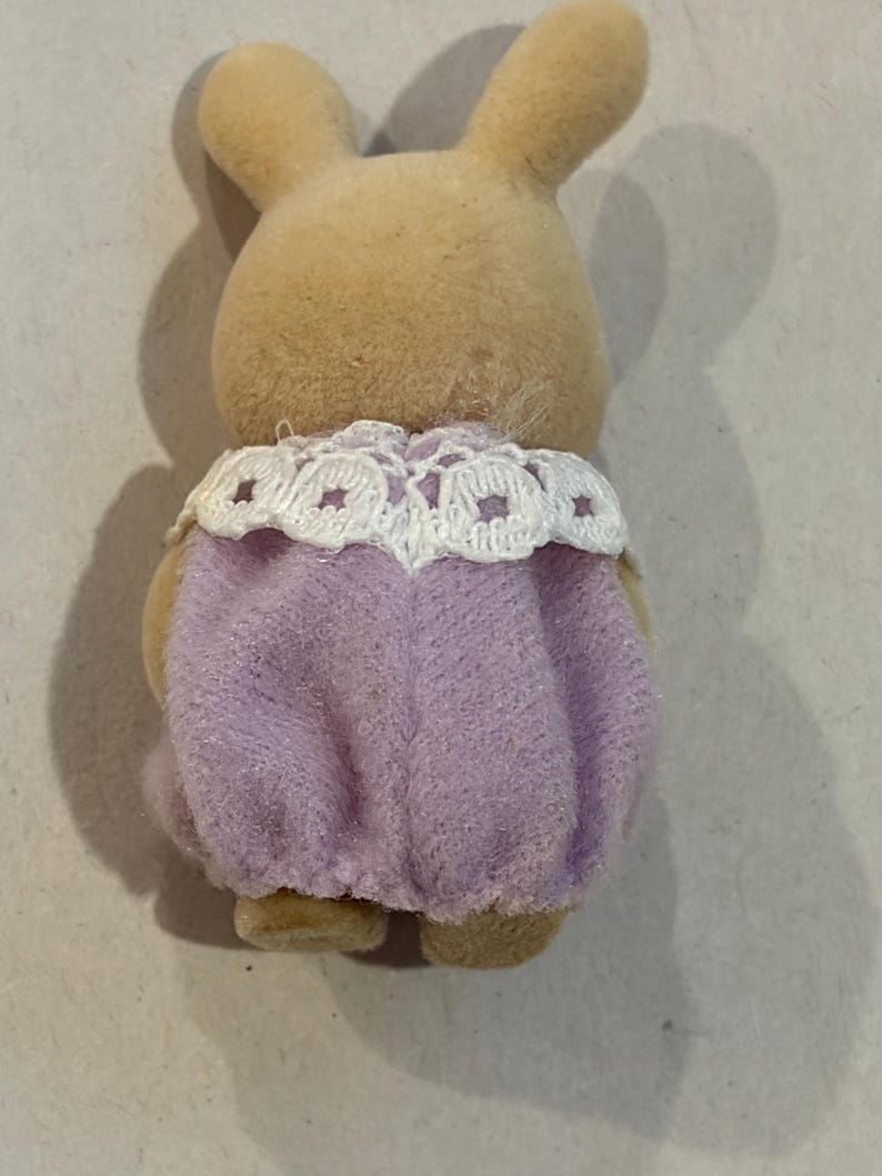 K&ouml;nnte beinhalten: Eine hellbraune Spielzeug-Kaninchenfigur, von hinten gesehen, mit einem lavendelfarbenen Outfit und einem wei&szlig;en Spitzenkragen. Das Kaninchen hat lange Ohren und eine weiche, flauschige Textur, typisch f&uuml;r Kinderspielzeug.