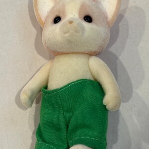 K&ouml;nnte beinhalten: Ein beflocktes Spielzeug-Hundefigur mit cremefarbenem K&ouml;rper und einem rosa-cremefarbenen Kopf. Der Hund tr&auml;gt eine gr&uuml;ne Hose. Das Spielzeug hat schwarze Augen und steht aufrecht.