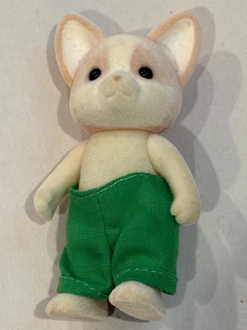 K&ouml;nnte beinhalten: Ein beflocktes Spielzeug-Hundefigur mit cremefarbenem K&ouml;rper und einem rosa-cremefarbenen Kopf. Der Hund tr&auml;gt eine gr&uuml;ne Hose. Das Spielzeug hat schwarze Augen und steht aufrecht.