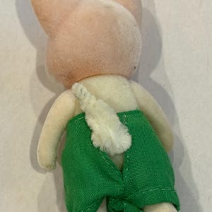 K&ouml;nnte beinhalten: Ein Vintage-Pl&uuml;schtier-Hase, von hinten gesehen. Der Hase hat einen hellrosa Kopf und Ohren, einen cremefarbenen K&ouml;rper und gr&uuml;ne Shorts. Ein flauschiger wei&szlig;er Schwanz ist sichtbar. Das Spielzeug stammt wahrscheinlich aus einer Kindersammlung.