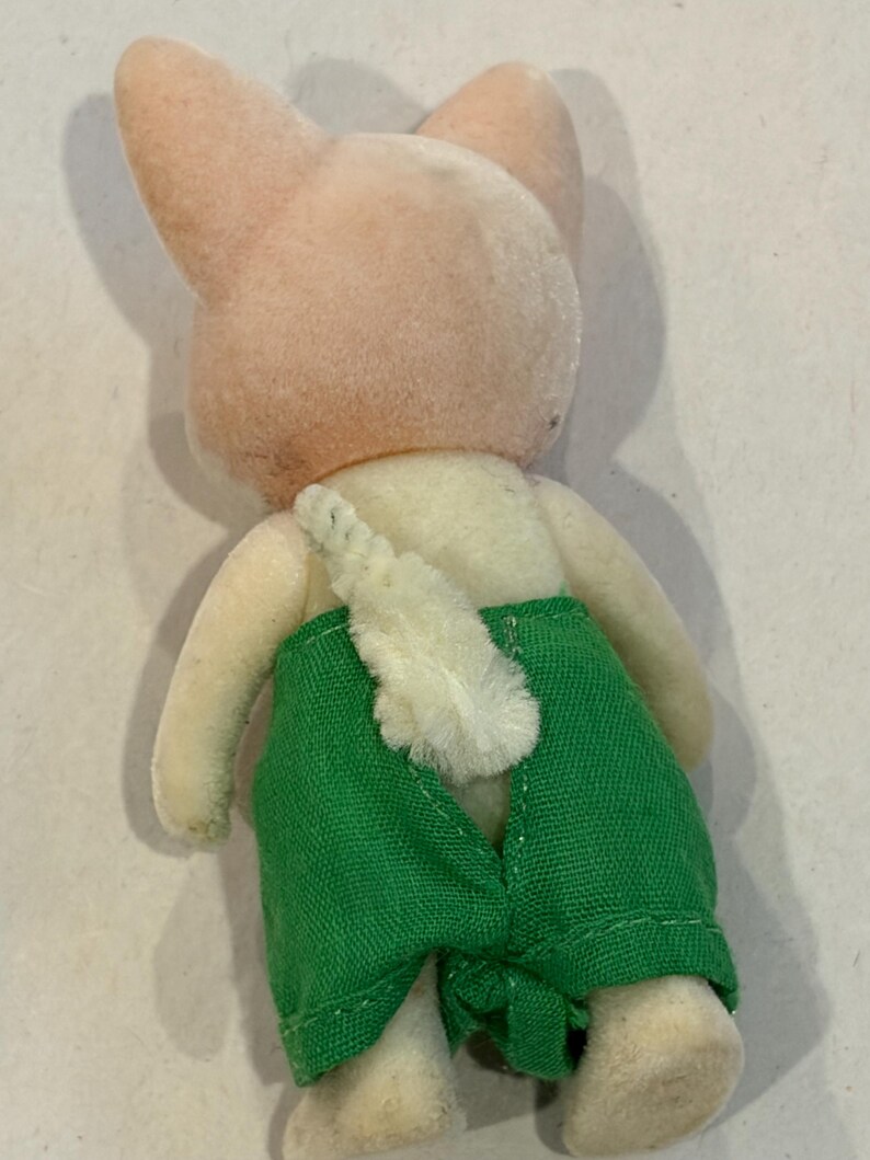K&ouml;nnte beinhalten: Ein Vintage-Pl&uuml;schtier-Hase, von hinten gesehen. Der Hase hat einen hellrosa Kopf und Ohren, einen cremefarbenen K&ouml;rper und gr&uuml;ne Shorts. Ein flauschiger wei&szlig;er Schwanz ist sichtbar. Das Spielzeug stammt wahrscheinlich aus einer Kindersammlung.