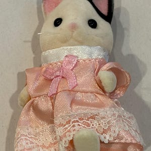 K&ouml;nnte beinhalten: Eine wei&szlig;-schwarze Katzenfigur, die ein rosa Kleid mit wei&szlig;er Spitzenborte und einer rosa Schleife tr&auml;gt. Die Katze hat schwarze Knopfaugen, rosa Ohren und eine rosa Nase. Das Kleid hat einen wei&szlig;en Kragen.