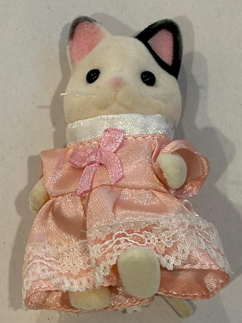 K&ouml;nnte beinhalten: Eine wei&szlig;-schwarze Katzenfigur, die ein rosa Kleid mit wei&szlig;er Spitzenborte und einer rosa Schleife tr&auml;gt. Die Katze hat schwarze Knopfaugen, rosa Ohren und eine rosa Nase. Das Kleid hat einen wei&szlig;en Kragen.