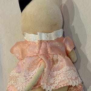 K&ouml;nnte beinhalten: Kleine Pl&uuml;schkatzenfigur von hinten gesehen. Die Katze hat einen cremefarbenen K&ouml;rper, schwarze Ohren und tr&auml;gt ein rosa Kleid mit wei&szlig;er Spitzenborte. Der Schwanz der Katze ist sichtbar.