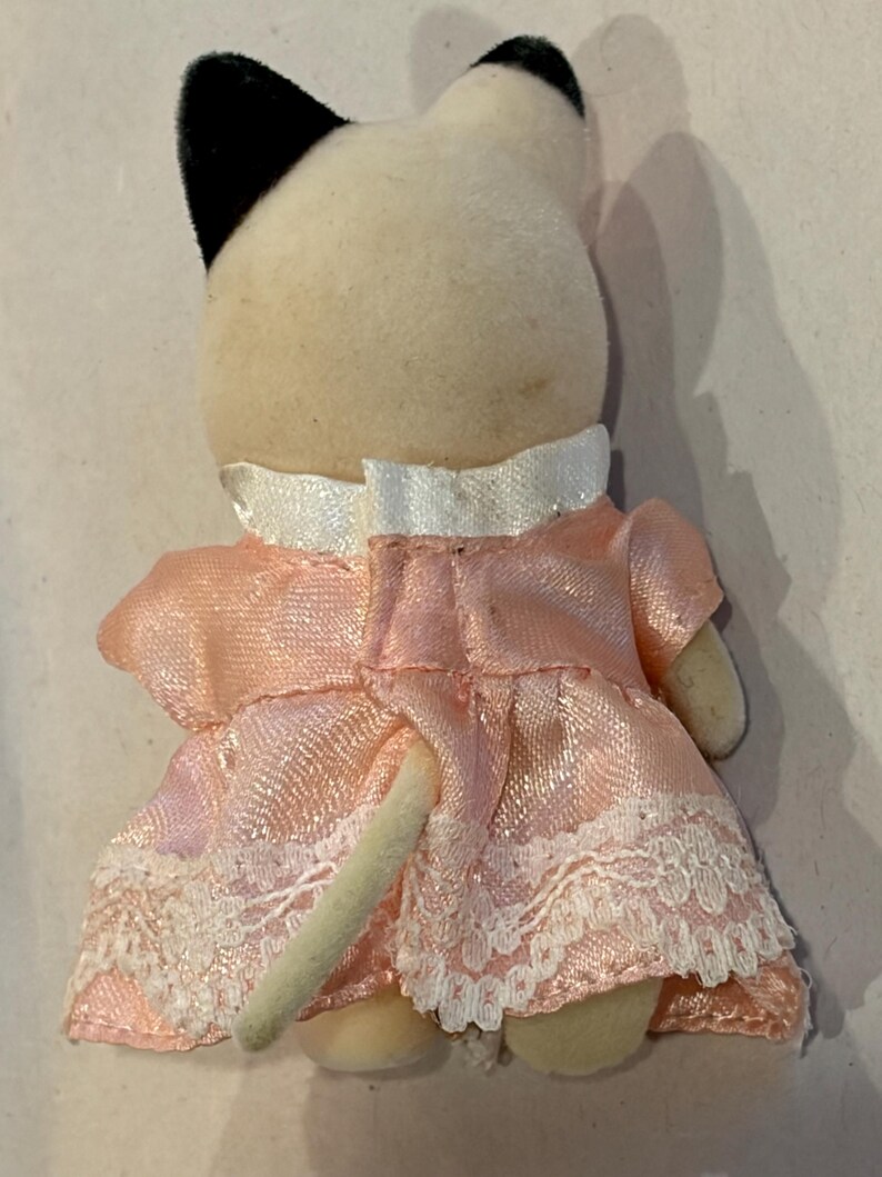 K&ouml;nnte beinhalten: Kleine Pl&uuml;schkatzenfigur von hinten gesehen. Die Katze hat einen cremefarbenen K&ouml;rper, schwarze Ohren und tr&auml;gt ein rosa Kleid mit wei&szlig;er Spitzenborte. Der Schwanz der Katze ist sichtbar.