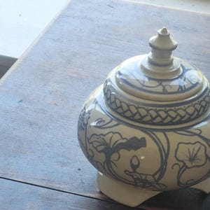 May include: A cream-colored ceramic jar with a matching lid, featuring a blue floral and vine pattern. The jar has a rounded body, a pointed finial on the lid, and stands on three small feet. The surface is smooth and glossy with a decorative braided band.