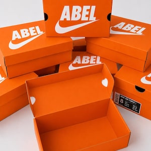 May include: A collection of bright orange rectangular cardboard shoe boxes stacked together. Each box features the word ABEL printed in bold white capital letters above a white swoosh graphic. One box is open, and another shows a size 8 label.