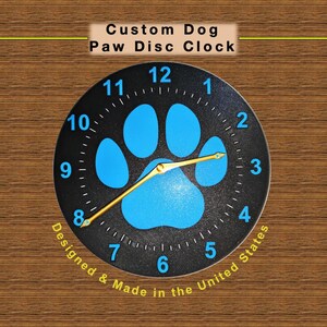 May include: A round wall clock with a black face featuring a blue paw print design. The clock has gold hands and white numbers. The text "Custom Dog Paw Disc Clock" is at the top. The words "Designed & Made in the United States" are at the bottom.