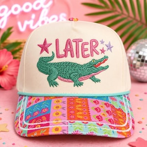 May include: A cream-colored baseball cap featuring an embroidered green alligator and the pink text LATER. The cap includes a light blue cord trim above the brim, which is decorated with a colorful patchwork pattern of flowers and geometric shapes.