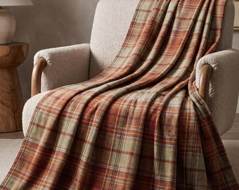 May include: A soft throw blanket featuring a classic plaid pattern in shades of burnt orange, muted sage green, and cream. The fabric drapes over a textured off-white armchair, showcasing a repeating grid of intersecting horizontal and vertical lines.
