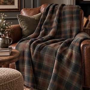 May include: A soft throw blanket featuring a classic plaid pattern in muted shades of forest green, charcoal gray, and burnt orange. The fabric has a smooth, uniform texture and is draped over a brown leather armchair, showcasing the woven design.
